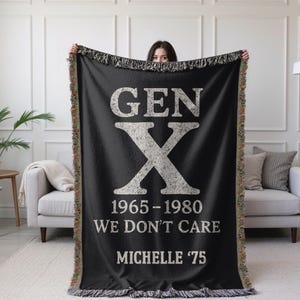 May include: Black throw blanket with white text reading "GEN X" and "1965-1980 WE DON'T CARE MICHELLE '75". The blanket has a fringed edge. The blanket is displayed in a living room setting. The blanket is being held up by a person.