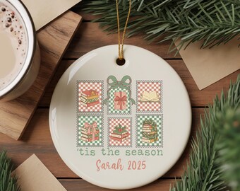 May include: A white ceramic Christmas ornament with a gold string. The ornament features a design of books and gifts in postage stamp style, with the text "'tis the season" and "Sarah 2025". The design includes green, red, and beige colors.