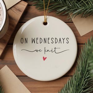 Könnte beinhalten: Runder, cremefarbener Keramikschmuck mit dem Text "ON WEDNESDAYS we knit" in schwarzer Schrift, einem kleinen roten Herz und einem goldenen Faden zum Aufhängen. Der Schmuck liegt auf einer Holzoberfläche mit Grün.
