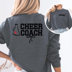 Puede incluir: Una sudadera gris oscuro con el texto "CHEER COACH" y un megáfono blanco y negro con un corazón rojo. La sudadera tiene un lazo debajo del texto.