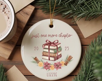 May include: A round, white ceramic ornament with a gold string for hanging. The ornament features a stack of books tied with a bow, the text "just one more chapter", and the name "LIZZIE". Decorative elements include cinnamon sticks, flowers, and orange slices.