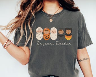 Könnte beinhalten: Dunkelgraues T-Shirt mit einem Halloween-Motiv. Das Design zeigt fünf Cartoonfiguren, die als Mumie, Kürbis, Geist, Fledermaus und Zuckermais verkleidet sind. Darunter steht der Schriftzug "Daycare Teacher".