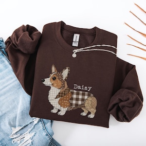 May include: A dark brown sweatshirt featuring a patchwork corgi dog design with the name "Daisy" in white. The corgi has a mix of patterns, including polka dots and plaid. A silver necklace with a small pendant is draped across the sweatshirt.