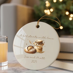 May include: A round, cream-colored ceramic ornament with a gold string for hanging. It features a watercolor illustration of bread and a jar of honey, with the text "it all starts with love", "Jess and Matt", and the year 2025.