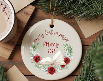 May include: A round, cream-colored ceramic ornament with gold string. The ornament features the text "easily lost in pages" and "Macy 2025" in a floral wreath design with red roses and an open book illustration.
