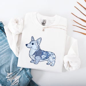 May include: White sweatshirt featuring a blue and white patterned Corgi dog design. The dog illustration incorporates floral and patchwork patterns. A silver necklace with a small pendant is draped across the sweatshirt. Blue jeans are visible in the background.