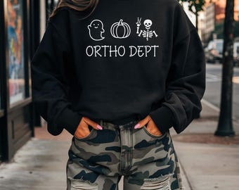 Peut inclure: Sweat-shirt noir avec un motif blanc d'un fantôme, d'une citrouille et d'un squelette avec le texte "ORTHO DEPT". La personne porte un short de camouflage.