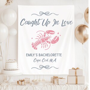 Puede incluir: Bandera de tela blanca con el texto "Caught Up In Love" y "Emily's Bachelorette Cape Cod, MA". Un gráfico de langosta rosa está centrado en la bandera. La bandera está decorada con lazos y estrellas.
