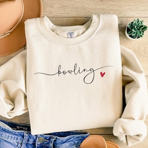 May include: Cream-colored sweatshirt with the word "bowling" in black script and a small red heart. The sweatshirt is folded, with a portion of blue jeans and a brown hat visible in the background. A small succulent plant is also visible.