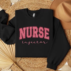 Könnte beinhalten: Schwarzer Pullover mit dem Text "NURSE Injector" in rosa Schrift. Der Text "NURSE" ist in einer großen, fetten Schrift mit einem Vintage-Look.