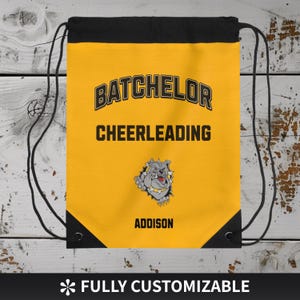 May include: A yellow drawstring bag with black accents and drawstrings. The bag features the text "BATCHELOR CHEERLEADING" in black and gold, along with a bulldog mascot and the name "ADDISON". The bottom of the bag reads "FULLY CUSTOMIZABLE".