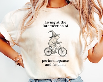 May include: A cream-coloured t-shirt with a humorous design. It shows a frog in a wizard hat on a bicycle, with the text: "Living at the intersection of perimenopause and fascism."
