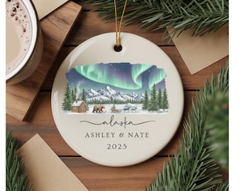 Puede incluir: Adorno navideño redondo de cerámica blanquecina con una cuerda dorada. Presenta una ilustración en acuarela de la aurora boreal de Alaska, montañas y una cabaña. El texto incluye "alaska", "ASHLEY & NATE" y "2025".