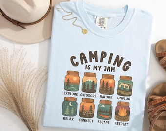 May include: Light blue t-shirt with the text "CAMPING IS MY JAM" and illustrations of camping scenes in mason jars. The shirt also features a hat, necklace, and sandals.