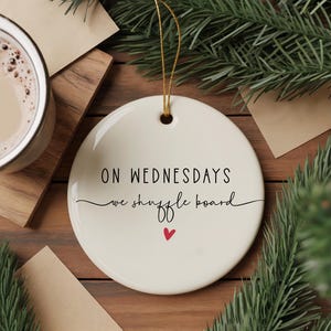 Pode incluir: Um ornamento redondo de cerâmica branca com o texto "ON WEDNESDAYS we shuffle board" em escrita preta. Um pequeno coração vermelho está abaixo do texto. O ornamento pende de um fio dourado.