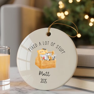 Puede incluir: Adorno navideño redondo de cerámica blanquecina con una cuerda dorada para colgar. El adorno presenta el texto "FIXED A LOT OF STUFF" y una ilustración en acuarela de una caja de herramientas. Debajo de la ilustración está el nombre "Matt" y el año "2025".