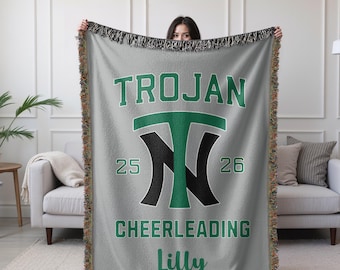 May include: A gray woven throw blanket with a fringed edge. The blanket features the text "TROJAN CHEERLEADING" in green and black, along with the name "Lilly". The design includes the numbers "25" and "26".