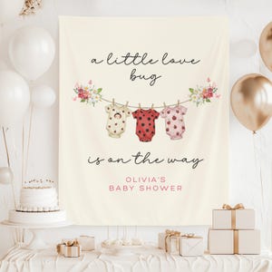 以下が含まれることがあります： 「a little love bug is on the way」の文字が入ったクリーム色のベビーシャワーバナー。てんとう虫のデザインのロンパースが3つ、物干しロープに吊るされています。バナーは花のアクセントと「Olivia's Baby Shower」の名前で飾られています。