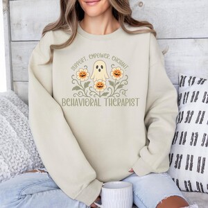 May include: Beige sweatshirt with a graphic featuring a ghost, flowers, and the words "SUPPORT. EMPOWER. ENCHANT. BEHAVIORAL THERAPIST" in a whimsical design. The sweatshirt is a neutral color and has long sleeves.