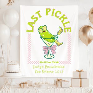 May include: A white bachelorette party backdrop with a pink and white checkered pattern. The backdrop features a cartoon pickle in a martini glass with the text "Last Pickle" in a green and pink font. The text "She'll Ever Tickle" is below the pickle, followed by "Emily's Bachelorette New Orleans 2023".