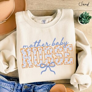 May include: A sand-coloured crewneck sweatshirt featuring the text "mother baby" in blue script above the word "NURSE" in large block letters. The block letters have a white background with a blue floral and orange pumpkin pattern.