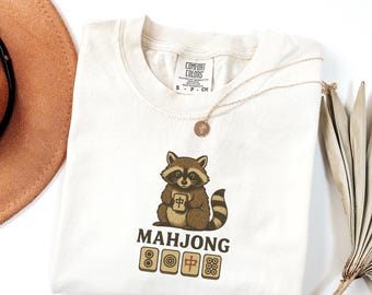 Könnte beinhalten: Cremefarbenes T-Shirt mit einem Waschbären, der einen Mahjong-Stein hält. Auf dem Shirt steht das Wort "MAHJONG" unter dem Waschbären, zusammen mit vier Mahjong-Steinen. Eine goldene Halskette und ein brauner Hut sind ebenfalls zu sehen.
