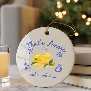 Może przedstawiać: Okrągła, kremowa ceramiczna ozdoba świąteczna z niebieskim napisem "That's Amore", ilustracją cytryny i imionami "John and Gia". Posiada złotą zawieszkę i rok 2025.