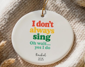 Może przedstawiać: Okrągła, biała ceramiczna ozdoba świąteczna z napisem "I don't always sing, Oh wait... yes I do" w kolorach czerwonym, żółtym, zielonym i turkusowym. Ozdoba zawiera imię "Rachel" i rok "2025". Dołączony jest złoty sznurek.