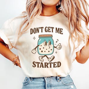 May include: Cream-colored t-shirt featuring a cartoon jar with a face, arms, and legs. The jar is light blue with a brown lid and black spots. The text "DON'T GET ME STARTED" is printed above and below the jar in black.