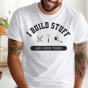 Op de afbeelding: Wit T-shirt met zwarte tekst die "I build stuff and I know things" luidt. De tekst is omgeven door een zwarte cirkel. In de cirkel bevinden zich drie gereedschappen: een gradenboog, een hamer en een meetlint.