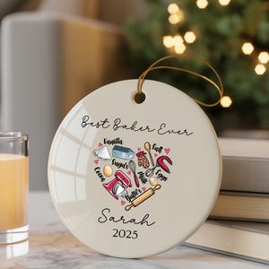 May include: A round, cream-colored ornament with a gold string for hanging. The ornament features the text "Best Baker Ever" and a heart-shaped design of baking ingredients and tools. The name "Sarah" and the year "2025" are also printed on the ornament.