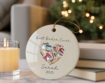 May include: A circular, cream-coloured ornament with a gold hanging string. It displays the text "Best Baker Ever" and a heart-shaped design of baking ingredients and tools. The name "Sarah" and the year "2025" are also printed on the ornament.