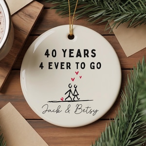 Op de afbeelding: Een rond wit keramieken ornament met de zwarte tekst 40 YEARS 4 EVER TO GO, een stokfiguurillustratie van twee lopende personen, drie kleine rode hartjes en de namen Jack & Betsy in zwart handschrift, opgehangen aan een gouden koordje.