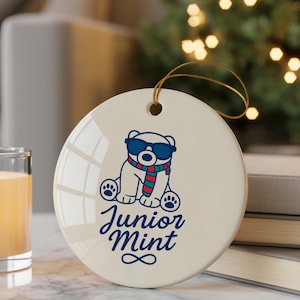 May include: A round, off-white ceramic ornament with a cartoon polar bear wearing sunglasses and a scarf. The text "Junior Mint" is written below the bear in a script font. A gold string is attached for hanging.