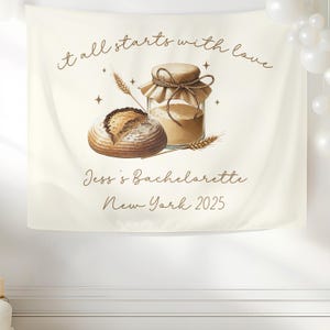 May include: Cream-colored fabric banner with the text "it all starts with love." A watercolor illustration depicts a loaf of bread, a jar of flour, and wheat stalks. The banner also reads "Jess's Bachelorette New York 2025."