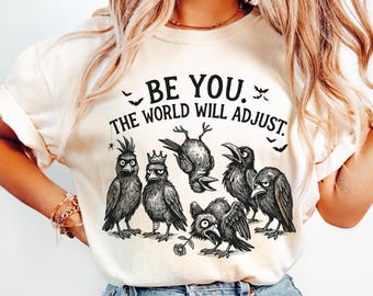 Pode incluir: Camiseta creme com um gráfico de seis corvos estilizados com várias expressões. O texto "BE YOU. THE WORLD WILL ADJUST." está impresso acima dos corvos, com pequenas ilustrações de morcegos e teias de aranha.