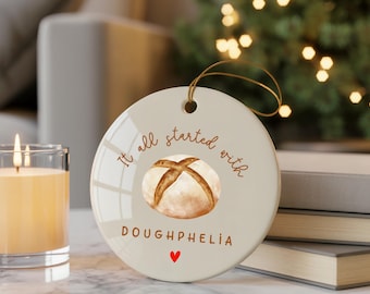 Puede incluir: Adorno navideño redondo de cerámica color crema con una cuerda dorada. Presenta una imagen de acuarela de una hogaza de pan y el texto "It all started with DOUGHPHELIA" con un corazón rojo. Una vela encendida y libros en el fondo.
