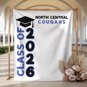 May include: White banner with the text "CLASS OF 2026" in blue and black, and "NORTH CENTRAL COUGARS" in black and blue. A black graduation cap is above the text. The banner is decorated with flowers and dried leaves.