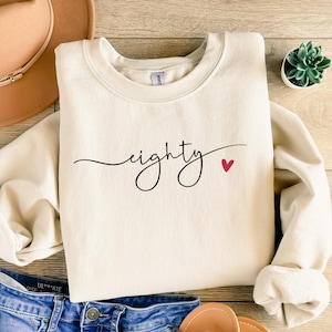 May include: Cream-colored sweatshirt with the word "eighty" in a cursive font and a small red heart. The sweatshirt is folded on a wooden surface, with a hat and jeans visible.