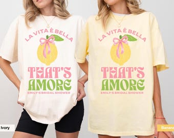 Peut inclure: Deux t-shirts blancs avec un motif de citron et le texte "La Vita è Bella That's Amore Emily's Bridal Shower". Un t-shirt est ivoire et l'autre est jaune banane.