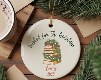May include: Round white ceramic Christmas ornament with a gold string. The ornament features a stack of books decorated with a garland and the text "booked for the holidays". Below the books is the name "Emily" and the year "2025".