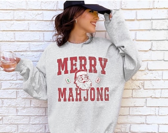 May include: A light gray sweatshirt with the words "MERRY MAHJONG" in red block letters. The design includes a Santa Claus graphic and Mahjong tiles. The sweatshirt is a festive holiday themed apparel item.