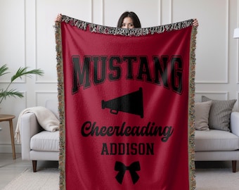 May include: A red throw blanket with black text and graphics. The word "MUSTANG" is at the top, with a megaphone graphic below it. The words "Cheerleading" and "ADDISON" are also displayed, along with a bow graphic.