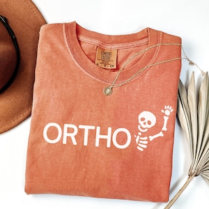 May include: An orange Comfort Colors t-shirt with the word "ORTHO" in white block letters and a white skeleton graphic waving. A gold necklace with a pendant and a brown hat are also visible.