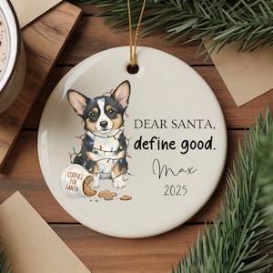 May include: A round ceramic Christmas ornament featuring a corgi wearing Christmas lights. The ornament has the text "DEAR SANTA, define good. Max 2025" and a small cookie jar that says "COOKIES FOR SANTA".