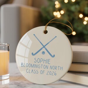Puede incluir: Adorno redondo de cerámica blanquecina con una cuerda dorada para colgar. Presenta un diseño azul de palo y disco de hockey, con el texto "SOPHIE BLOOMINGTON NORTH CLASS OF 2026" en azul. Una vela encendida y libros en el fondo.