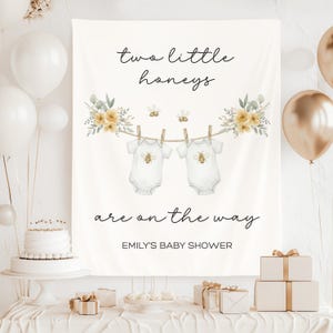May include: A white baby shower banner with the text "two little honeys are on the way." The banner features watercolour illustrations of baby onesies with bee designs hanging on a clothesline, and floral accents. The banner is for Emily's baby shower.