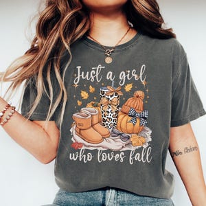 May include: Dark gray t-shirt with the text "Just a girl who loves fall." The graphic includes fall-themed illustrations: a pumpkin, boots, a tumbler, and autumn leaves. The tumbler has a leopard print design.