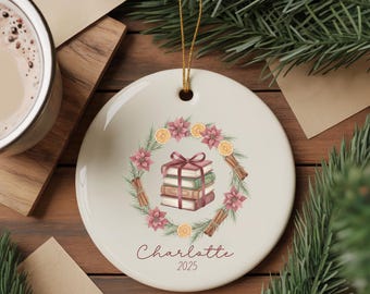May include: A round, white ceramic Christmas ornament with a gold string. The ornament features a watercolor design of a stack of books tied with a bow, surrounded by a wreath of flowers, orange slices, and cinnamon sticks. The name "Charlotte" and the year "2025" are written on the ornament.