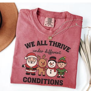 May include: A maroon Comfort Colors t-shirt with the text "WE ALL THRIVE under different CONDITIONS" and cartoon illustrations of Santa, a reindeer, a snowman, and an elf. A silver necklace with a pendant is draped across the shirt.